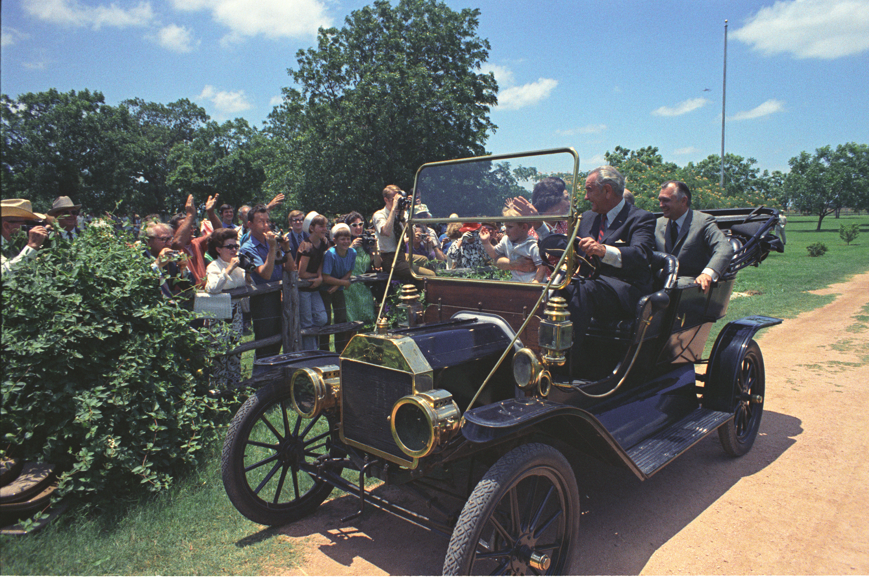 President Lyndon B. Johnson driving his Model-T Ford down a dirt road, waving to crowd. Lady Bird Johnson, grandson Lyn Nugent and others ride along.